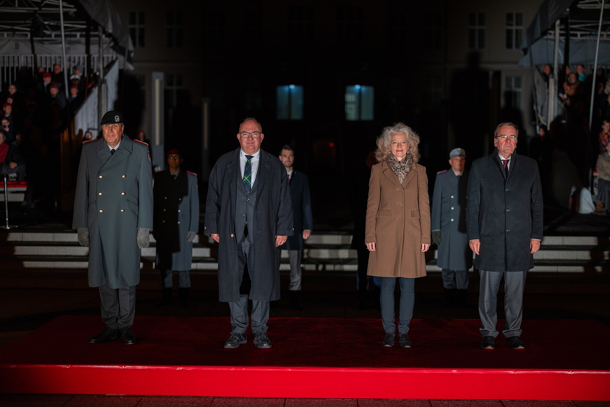 Außenaufnahme bei Dunkelheit. Im Hintergrund sitzen Zuschauende auf Tribünen, im Vordergrund auf einem roten Podest stehen von links nach rechts Generalleutnant Laubenthal in Uniform, in zivil Präsident Beckmann, Präsidentin Kern und Verteidigungsminister Pistorius