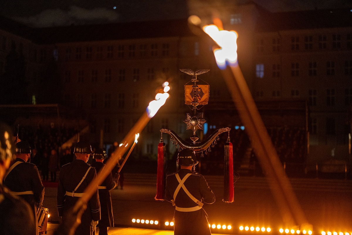 Außenaufnahme bei Dunkelheit: Im Hintergrund ist ein Gebäude zu sehen. Der Blick von hinten zeigt das Musikkorps sowie brennende Fackeln