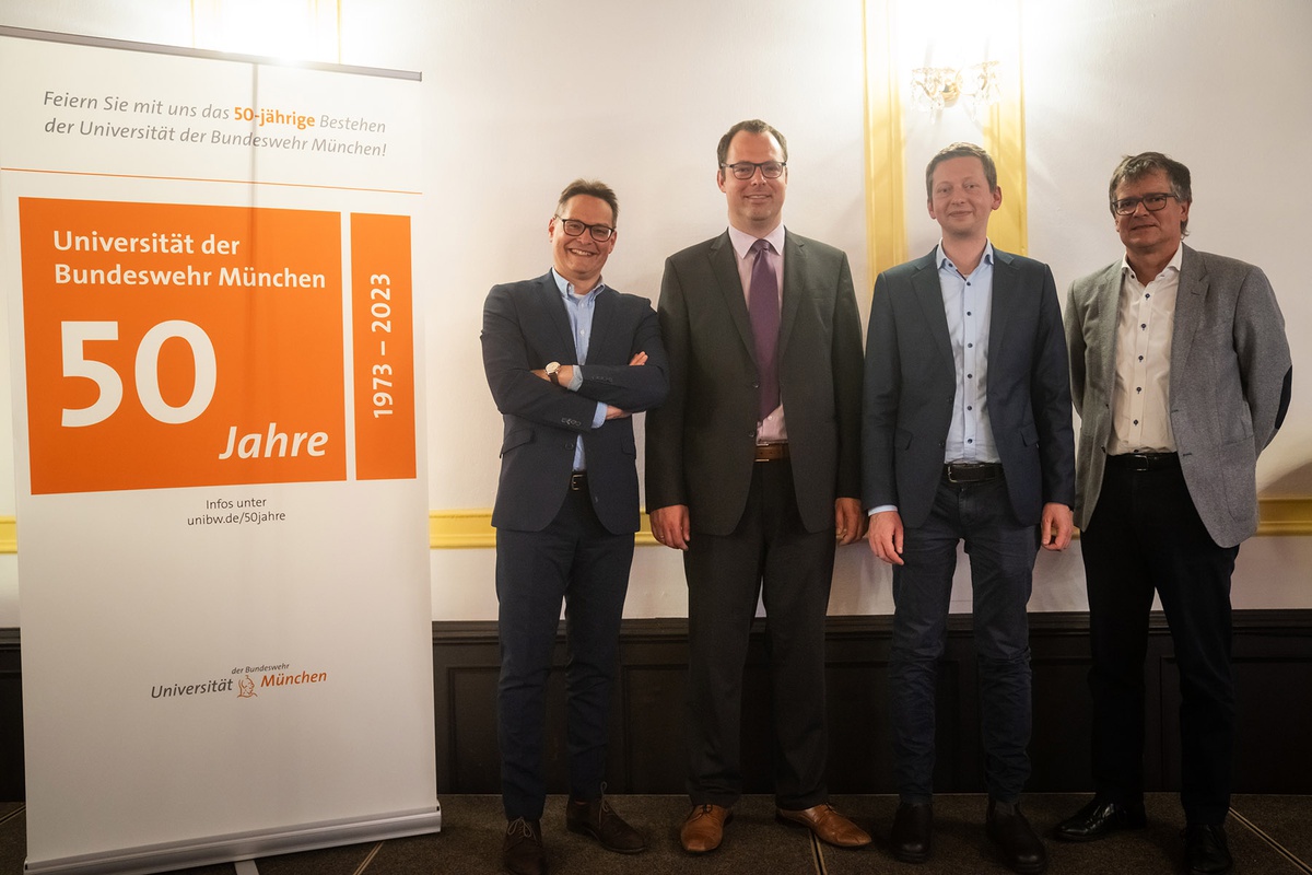 Links im Bild steht ein Banner mit dem Logo zum 50-jährigen Jubiläum der Universität, daneben stehen vier Männer in formeller Kleidung, der linke hat die Arme vor der Brust verschränkt, der rechte hinter dem Rücken, alle blicken freundlich in die Kamera.