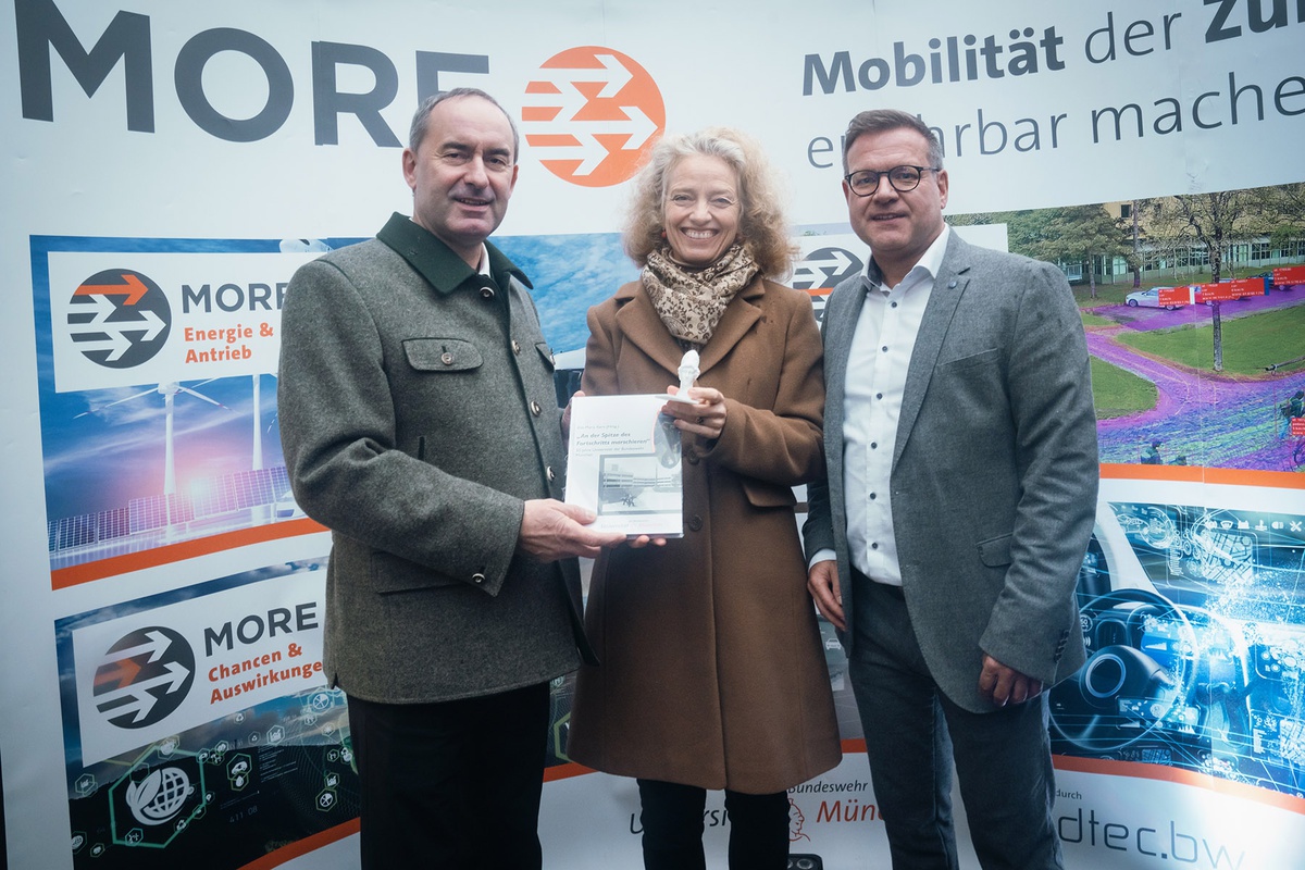 Gruppenfoto: links Staatsminister Aiwanger, in der Mitte Präsidentin Kern, rechts Prof. Trapp. Die drei stehen vor einer Fotowand des Projekts MORE. Präsidentin Kern überreicht Staatsminister Aiwanger die Athene sowie die Festschrift.