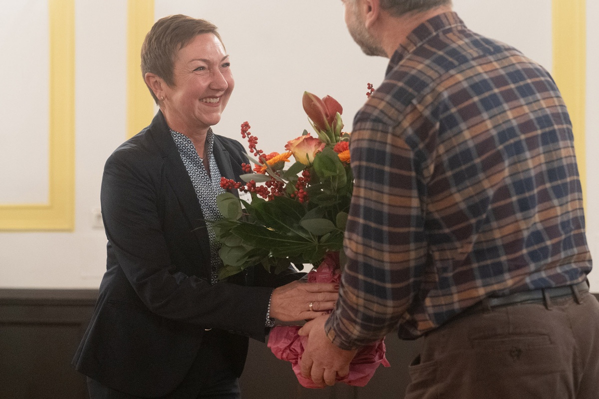 Dr. Matzner-Vogel steht links im Bild und nimmt lächeld einen großen Blumenstrauß entgegen, der ihr von einem Herren, der rechts im Bild von hinten zu sehen ist, überreicht wird
