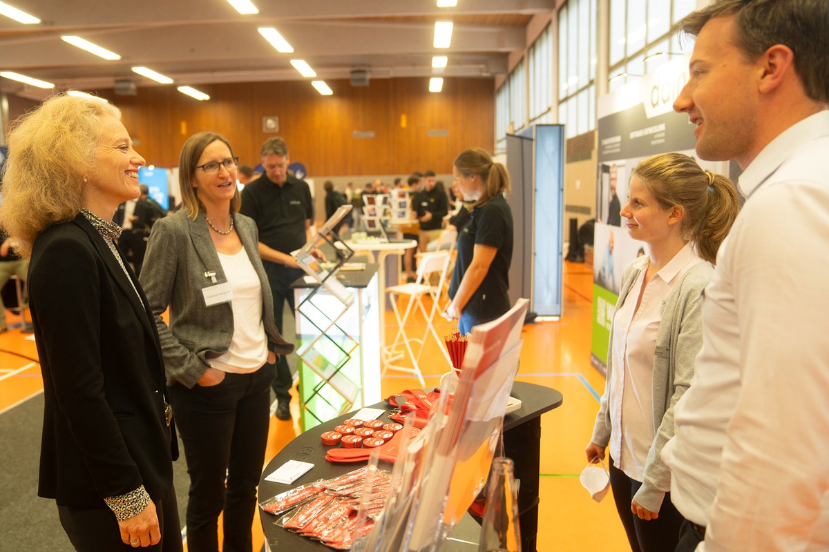 Ein Stand in der Sporthalle, in der die Veranstaltung stattfand. In der Bildmitte der Stand eines Unternehmens, rechts davon stehen eine Mitarbeiterin und ein Mitarbeiter, ihnen gegenüber stehen links im Bild Prof. Kern und Frau Borghoff vom Career Service. Die vier Personen befinden sich mitten im Gespräch.