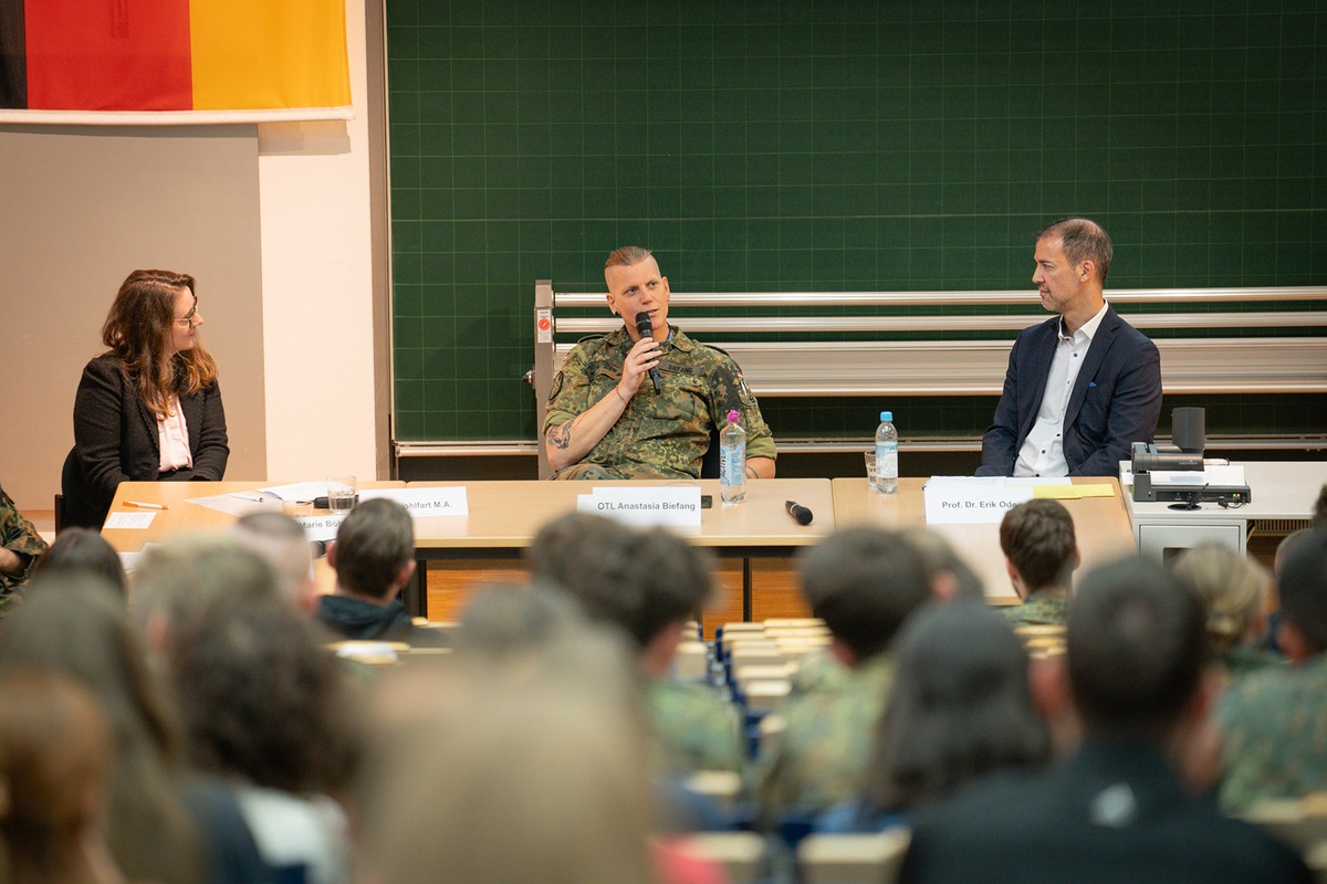 Im Bildvordergrund sieht man die Hinterköpfe der Studierenden, die nach vorne zu den Tischen schauen, an denen drei Personen sitzen, in der Mitte Obersetleutnant Biefang, die zum Publikum spricht