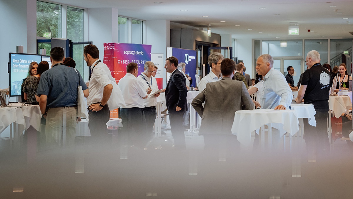 Mehrere Stehtische, an denen Menschen in Business-Kleidung stehen und sich unterhalten. Im Hintergrund sieht man Bildschirme und Roll-ups unterschiedlicher Firmen, beispielsweise der Fimra Spora Steria oder Airbus Defence sind zu lesen