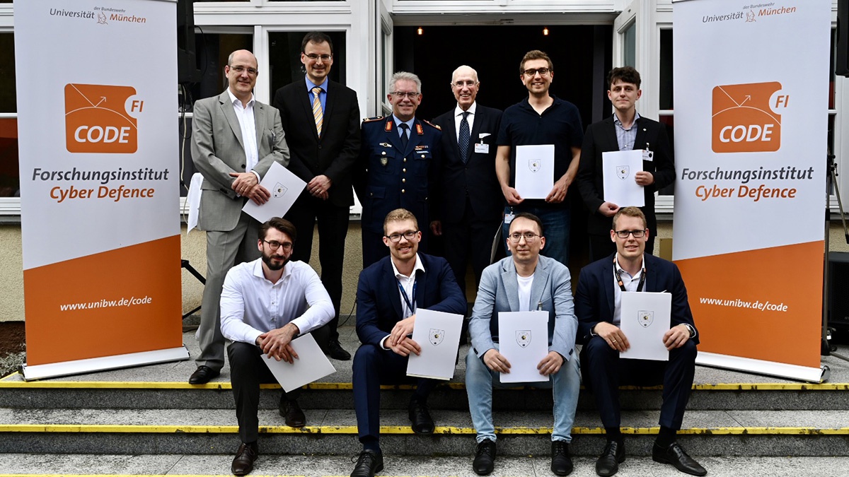 Links und rechts im Bild sind zwei Roll-ups des Forschungsinstituts Cyber Defence mit dessen Logo zu sehen, dazwischen befindet sich eine Gruppe Männer, die auf den Stufen steht (hintere Reihe) bzw. sitzt. Von den zehn Männern halten sieben etwas, das wie eine Urkunde aussieht, in den Händen. In der hinteren Reihe steht zwischen den Männern in Businesskleidung ein Brigadegeneral in Uniform 