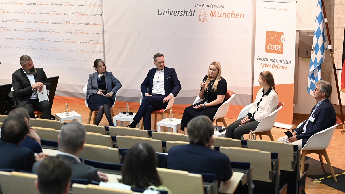 Vor einer Wand mit dem Signet der Universität und einem Roll-up mit dem Logo des Forschungsinstituts CODE sitzen frontal zum Publikum im Hörsaal drei Frauen und drei Männer nebeneinander. Eine der Frauen spricht in ein Mikrofon und zum Publikum, das auf dem Foto von hinten zu sehen ist.