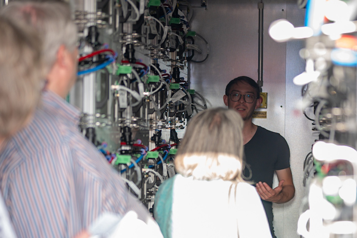 Blick von außen in einen Container, es sieht beengt aus. Das Bild wurde an den Zuhörenden vorbei fotografiert, die zum Projektmitarbeiter schauen. Dieser steht mit Blick in die Kamera im Container. Er ist umgeben von technischem Gerät, man kann bunte Kabel erkennen.