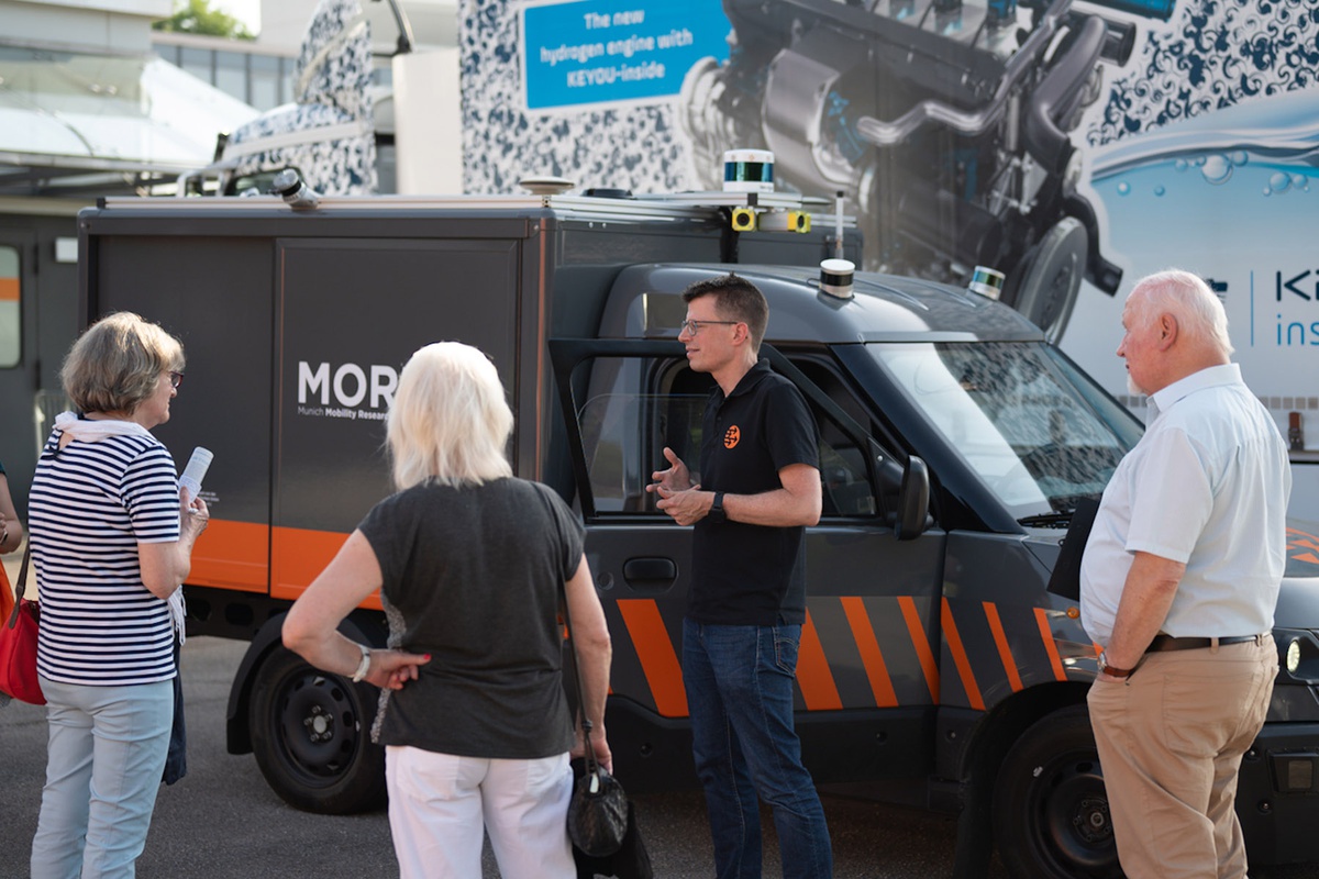 Ein Mitarbeiter des Projekts steht vor einem Auto, das wir ein Mini-LKW aussieht. Er spricht zu drei Personen, die vor ihm stehen und das Auto anschauen. Auf dem Auto kann man einige technische Gerätschaften erkennen, die auf dem Dach angebracht sind.