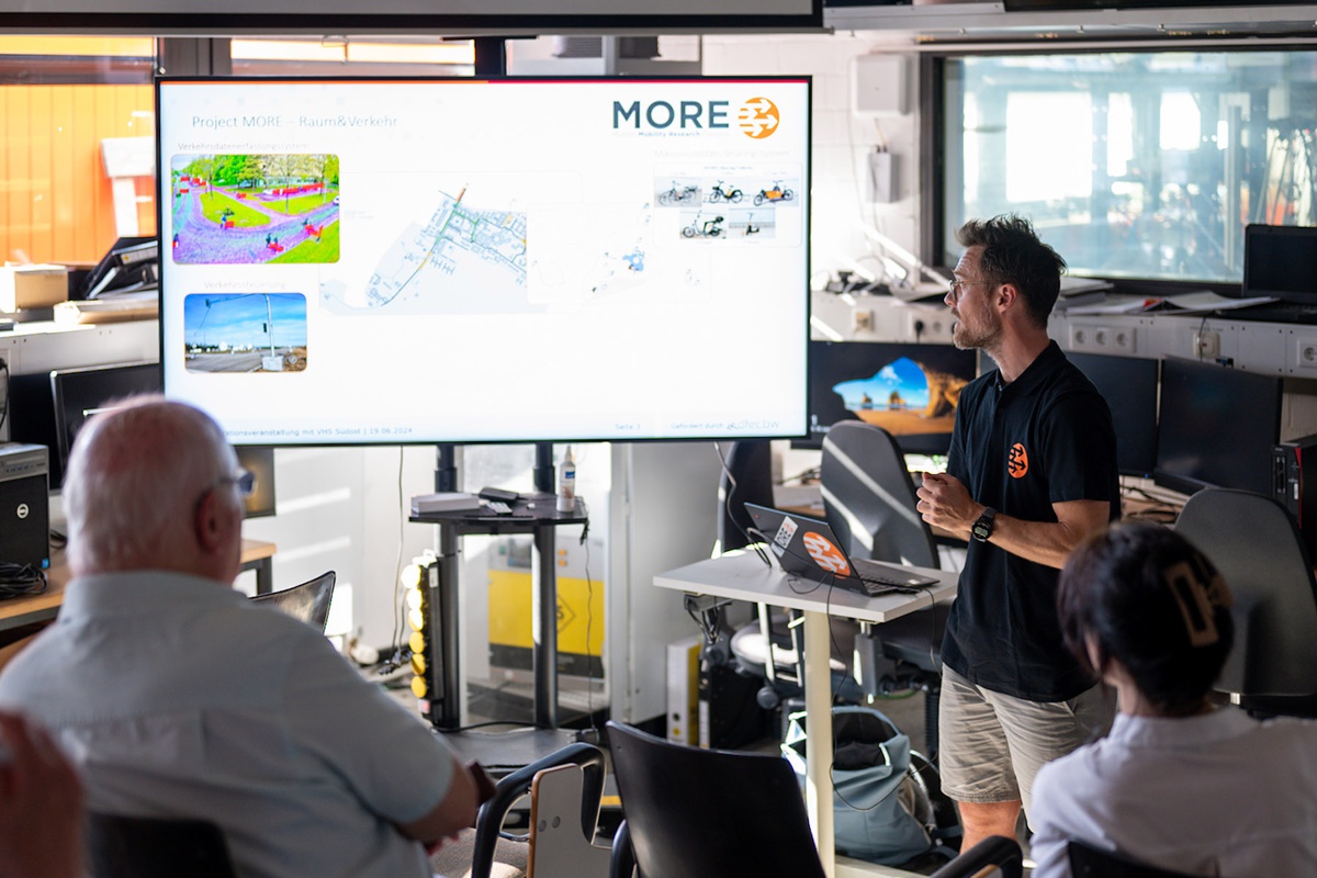 Ein Mitarbeiter des Projekts steht in einer Laborumgebung vor einem Bildschirm, auf den er schaut, während er spricht. Die Zuhörenden blicken ebenfalls auf den Monitor. Auf dem Monitor ist die Überschrift "Projekt MORE. Raum & Verkehr" zu erkenne. Darunter eine weitere Überschrift: Verkehrsdatenerfassungssystem. Darunter zwei Bilder, die Straßen und Wege auf dem Campus zeigen, daneben eine Karte des Campus und daneben Bilder der MORE-Mietfahrräder und -roller.