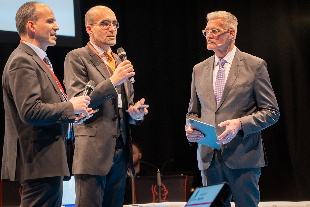 Links im Bild ein Mann im Anzug (Prof. Meurer), in der Mitte ein weiterer Mann im Anzug, der gerade in ein Mikrofon spricht (Prof. Pany) und rechts ein Mann im Anzug (Herr Kruesken), der Moderationskarten in der Hand hält und in Richtung der beiden anderen Männer blickt. 