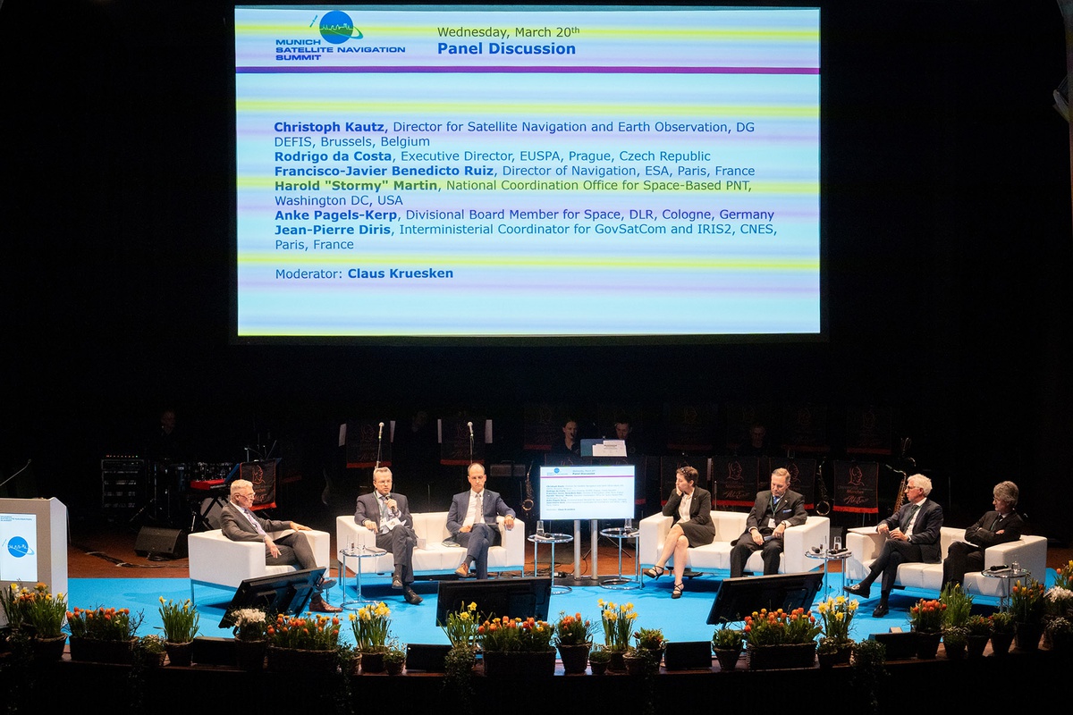 Blick auf die Bühne. Die Leinwand im Hintergrund zeigt eine Folie mit dem Logo des Summits. Zusätzlicher Text: "Wednesday, March 20th. Panel Discussion" sowie die Namen und Funktionen der Teilnehmenden. Auf hellen Sofas und Sesseln sitzen nebeneinander Moderator Kruesken und die Teilnehmenden Christoph Kautz, Rodrigo da Costa, Anke Pagels-Kerp, Harold Martin, Francisco-Javier Benedicto Ruiz und Jean-Pierre Diris