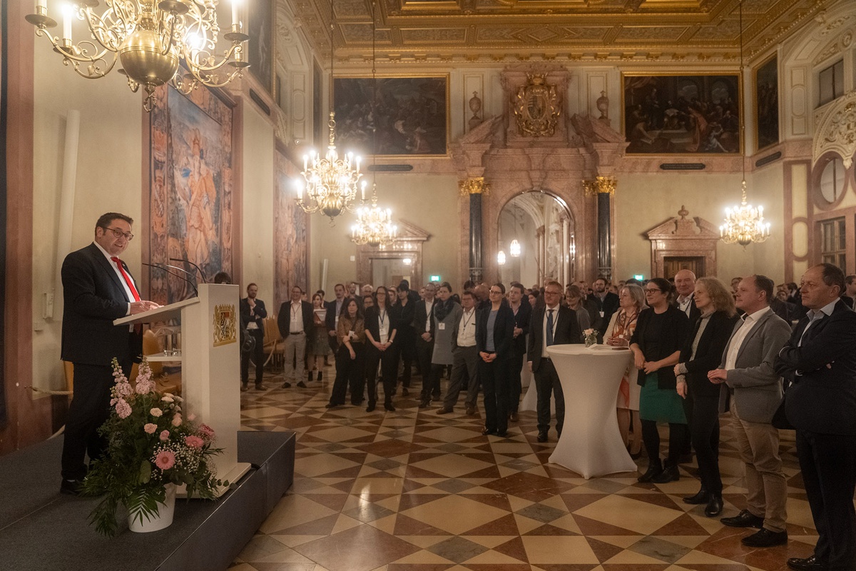 Blick in den Kaisersaal der Residenz. An den Wänden hängen Gemälde, die Decke ist mit goldfarbenem Stuck verziert, von der Decke hängen Kronleuchter. Auf einem kleinen Podest steht links im Bild Staatssekretär Tobias Gotthardt am Rednerpult und begrüßt die Anwesenden, die mit etwas Abstand um das Rednerpult herumgruppiert stehen