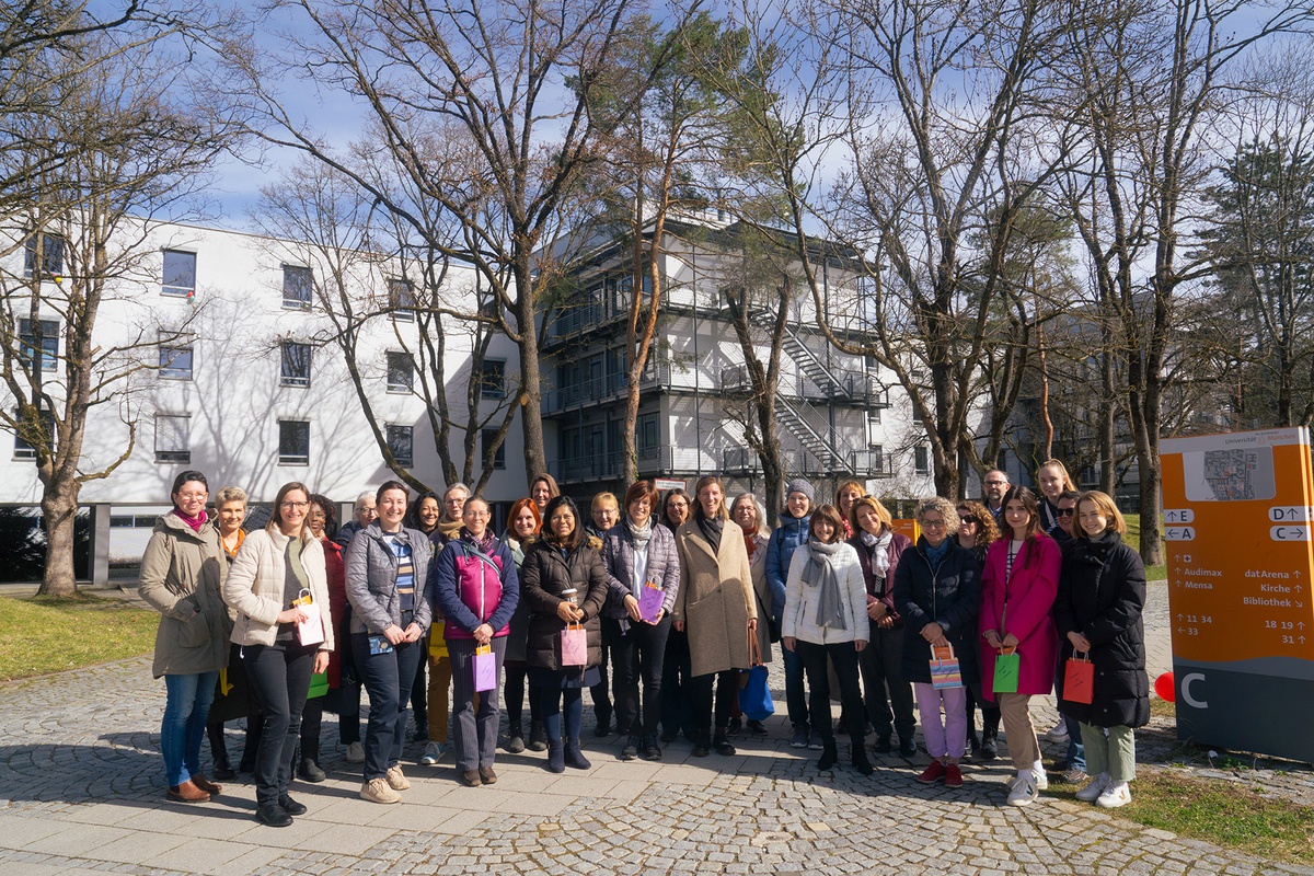 Gruppenfoto: eine Gruppe von ca. 25 Frauen und einem Mann steht auf dem Campus, im Hintergrund sind Bäume und ein Gebäude zu sehen. Die Sonne scheint, alle halten kleine bunte Tüten in der Hand.