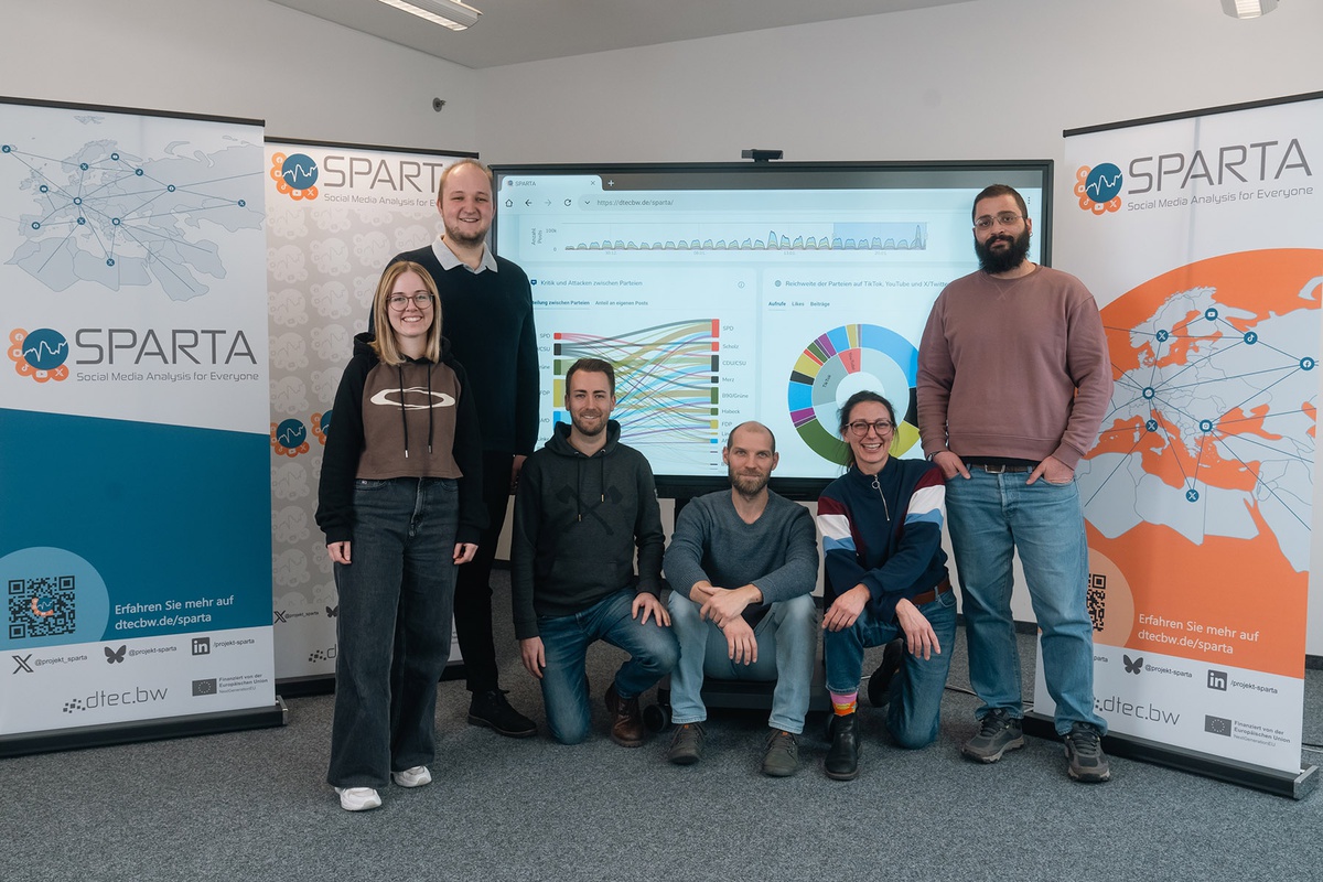 Gruppenfoto mit zwei Frauen und vier Männern, die vor Bannern des Projekts SPARTA stehen bzw. knien, direkt hinter der Gruppe zeigt ein großer Bildschirm verschiedene Diagramme