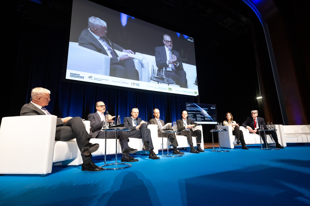 Sieben PErsonen sitzen auf weißen Sofas und sind einander zugewendet. ein Herr hat ein Mikro in der Hand und spricht.