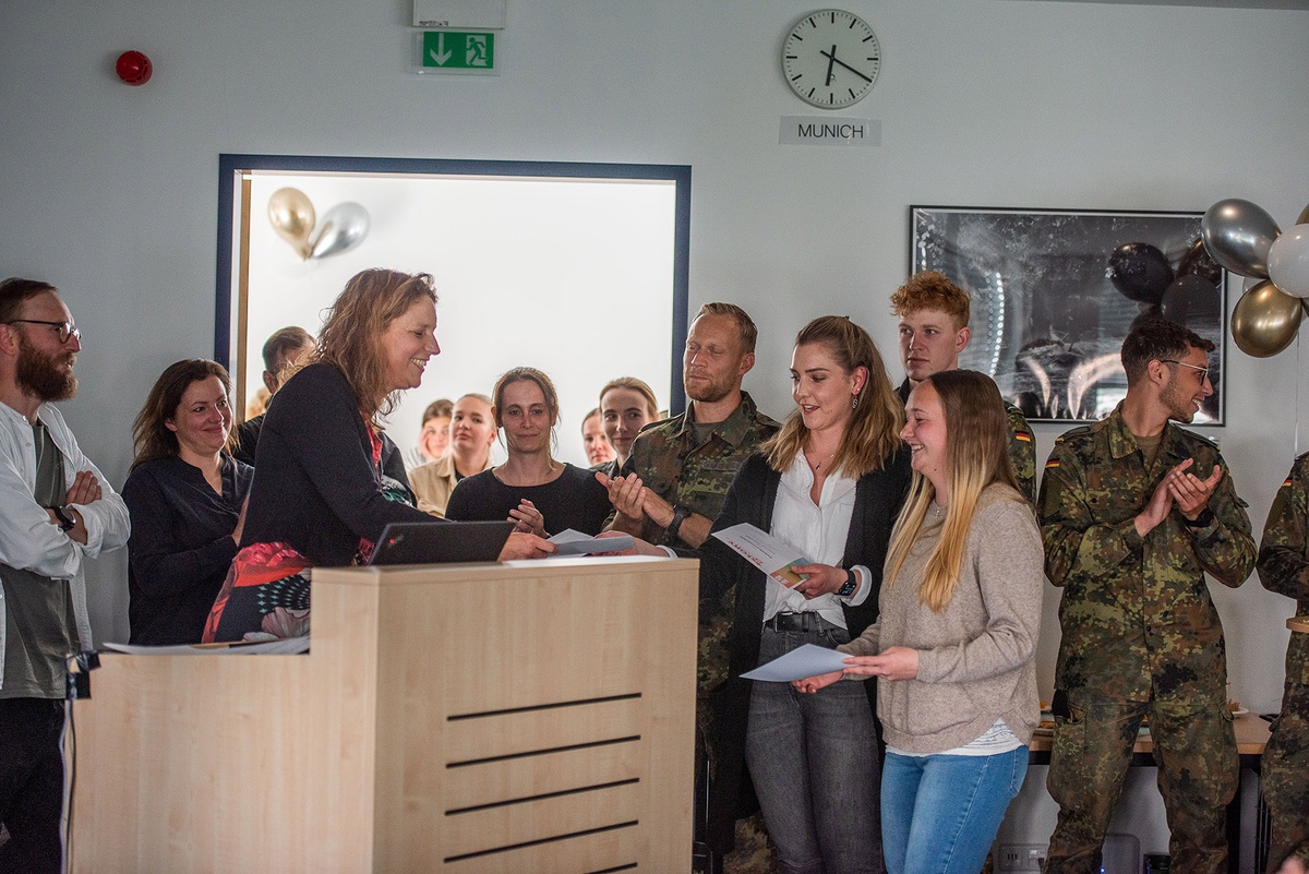 Die Professorin steht links im Bild hinter dem Rednerpult und überreicht den rechts davon stehenden Studentinnen eine Urkunde oder ähnliches. Im Hintergrund stehen Lehrpersonen sowie Studierende, einige von ihnen tragen Uniform.