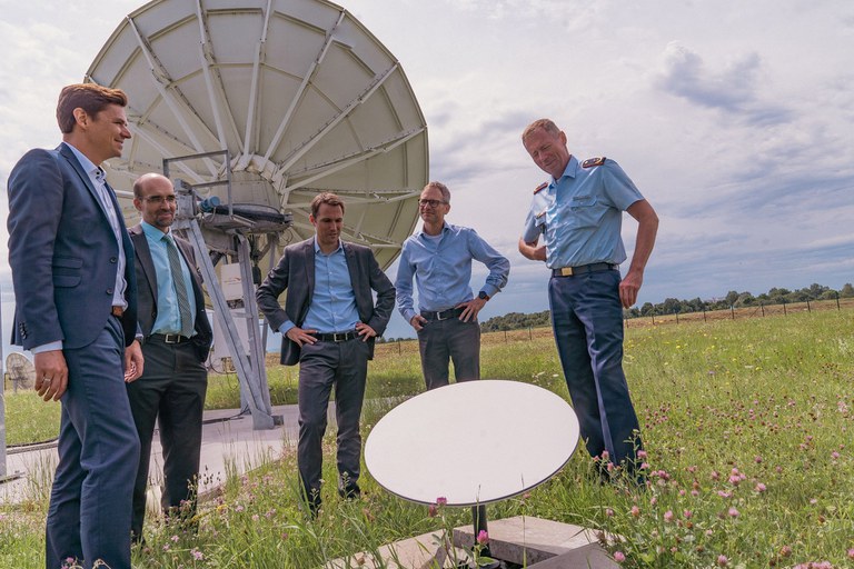 Eine Gruppe von Männern auf einer Wiese mit einer Antenne im Hintergrund und einem kleineren Gerät im Vordergrund.