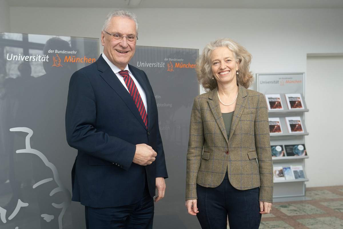Links im Bild innenminister Herrmann, rechts im Bild Präsidentin Prof. Kern. Die beiden stehen im Foyer vor einem grauen Hitnergrund mit dem Universitätslogo und der Athene.