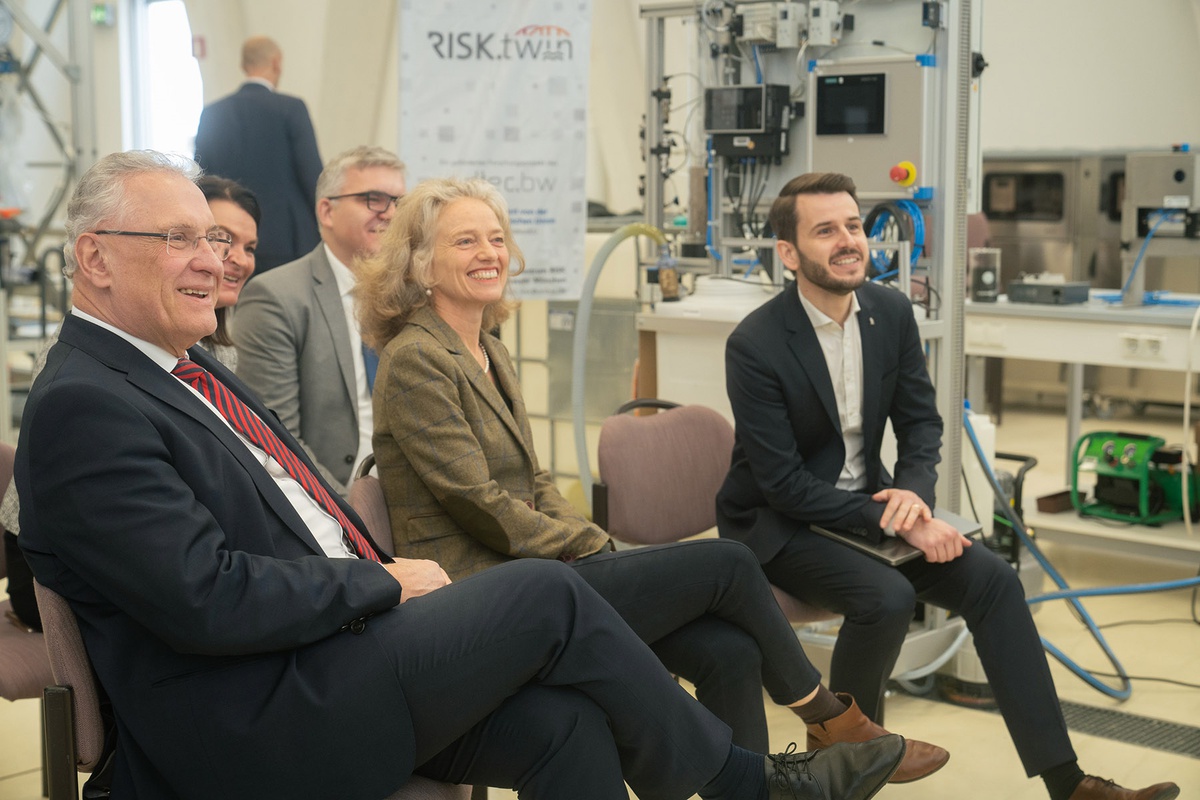 In einem Raum, in dessen Hintergrund der technische Gerätschaften zu sehen sind, befinden sich links im Bild mehrere Stühle, auf denen unter anderem links im Bild Innenminister Herrmann sitzt, danaben Prof. Kern und rechts Prof. Popp. Alle haben den Blick nach rechts gewandt und schauen auf etwas, das sich außerhalb des Bildes befindet.