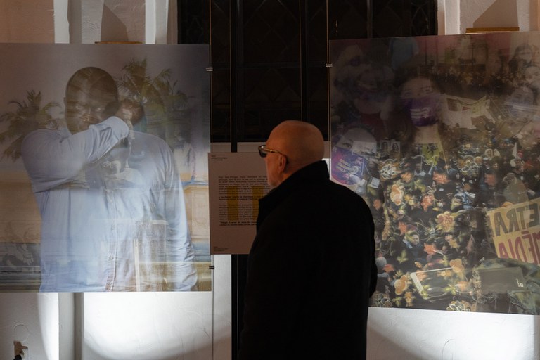 Zwei großformatige Bilder, davor steht – von hinten zu sehen – ein Mann und betrachtet eines davon, zu sehen ist eine Person of Color in hellem Hemd, im Hintergrund sind Palmen zu sehen. Der Mann auf dem Bild hält sich den angewinkelten Arm vor einen Teil seines Gesichts. Das andere Bild zeigt Menschen mit Spruchbändern, vermutlich eine Demonstration.