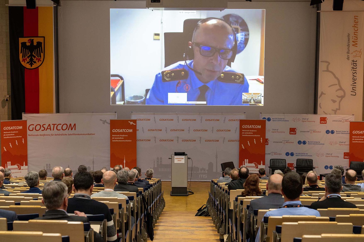 Audimax von hinten nach vorne fotografiert: auf einer großen Leinwand hinter dem Rednerpult ist Generalmajor Setzer zu sehen, der per Video zugeschaltet war.