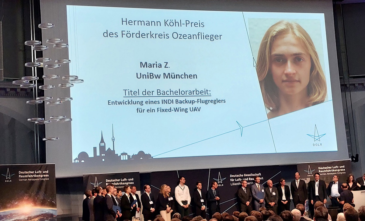 Von hinten nach vorne fotografiert: auf einer großen Leinwand ist das Bild von Maria Z. eingeblendet, daneben steht der Titel ihrer Arbeit.