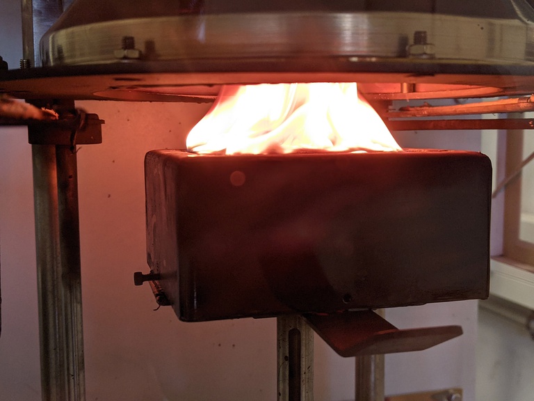 Auf einer Halterung ist ein großer Block befestigt, auf dem eine Materialprobe brennt. Darüber befindet sich eine runde Öffnung.