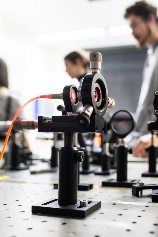 im Vordergrund steht ein Laborgerät mit Glasfaserkabel und Linse, welches zusammen mit einem roten Laser verwendet wird. Im Hintergrund sind weitere Gerätschaften, sowie drei Personen, jedoch ist hier alles verschwommen abgebildet.