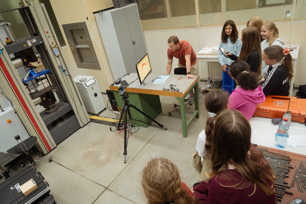 Eine Gruppe von Mädchen ist in einem Labor zu sehen, gemeinsam mit einem wissenschaftlichen Mitarbeiter. Auf der linken Seite befindet sich eine selber gebastelte Papierbrücke, die zum Testen in einer Maschine eingespannt ist.