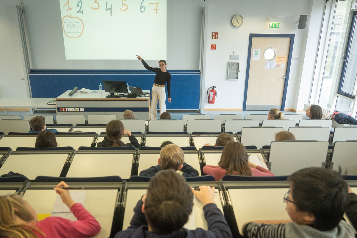 Im Hörsaal von hinten nach vorne fotografiert. In den Sitzreihen sitzen mehrere Kinder und schauen nach vorne, wo Prof. Riedl steht und den Kindern etwas erklärt, während sie auf eine Leinwand neben sich zeigt. Dort sind die Zahlen 1 bis 7 zu lesen.