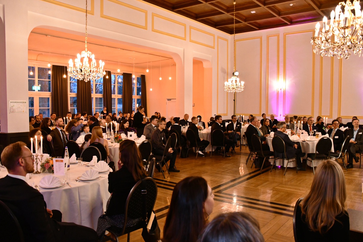 Blick in einen Saal mit Kronleuchtern, in dem festlich gedeckte Tische stehen, an denen die Absolventinnen und Absolventen mit ihren Familien sitzen