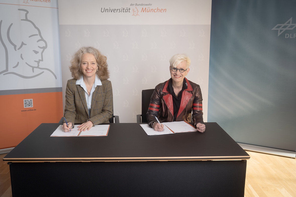 Prof. Kern (links) und Prof. Kaysser-Pyzalla sitzen an einem Tisch, beide haben Dokumente vor sich liegen. Die beiden Frauen haben je einen Stift in der Hand und schauen lächelnd in die Kamera. Im Hintergrund sind die Logos der Universität und des DLRs zu sehen.