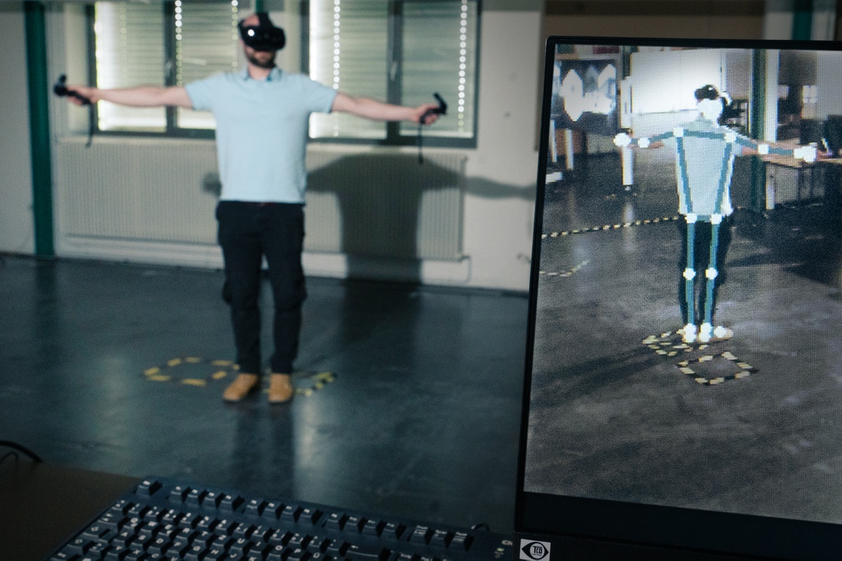 Im Hintergrund steht eine Person mit VR-Brille und Controllern in der Hand. Auf einem Bildschirm im Vordergrund ist eine graphische Darstellung der Person.