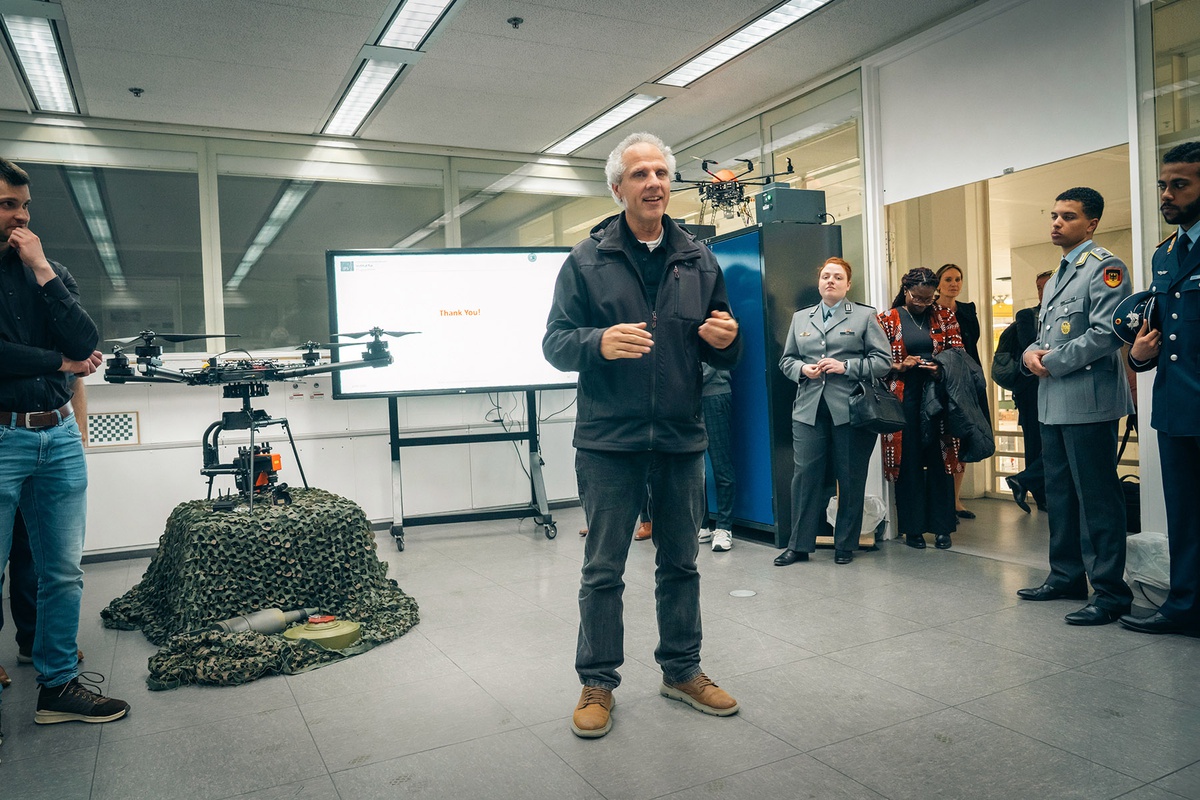 Prof. Stütz steht in seinem Labor, hinter ihm ist eine Srohne zu sehen. Die Gäste, zu denen er spricht, stehen an den Seiten