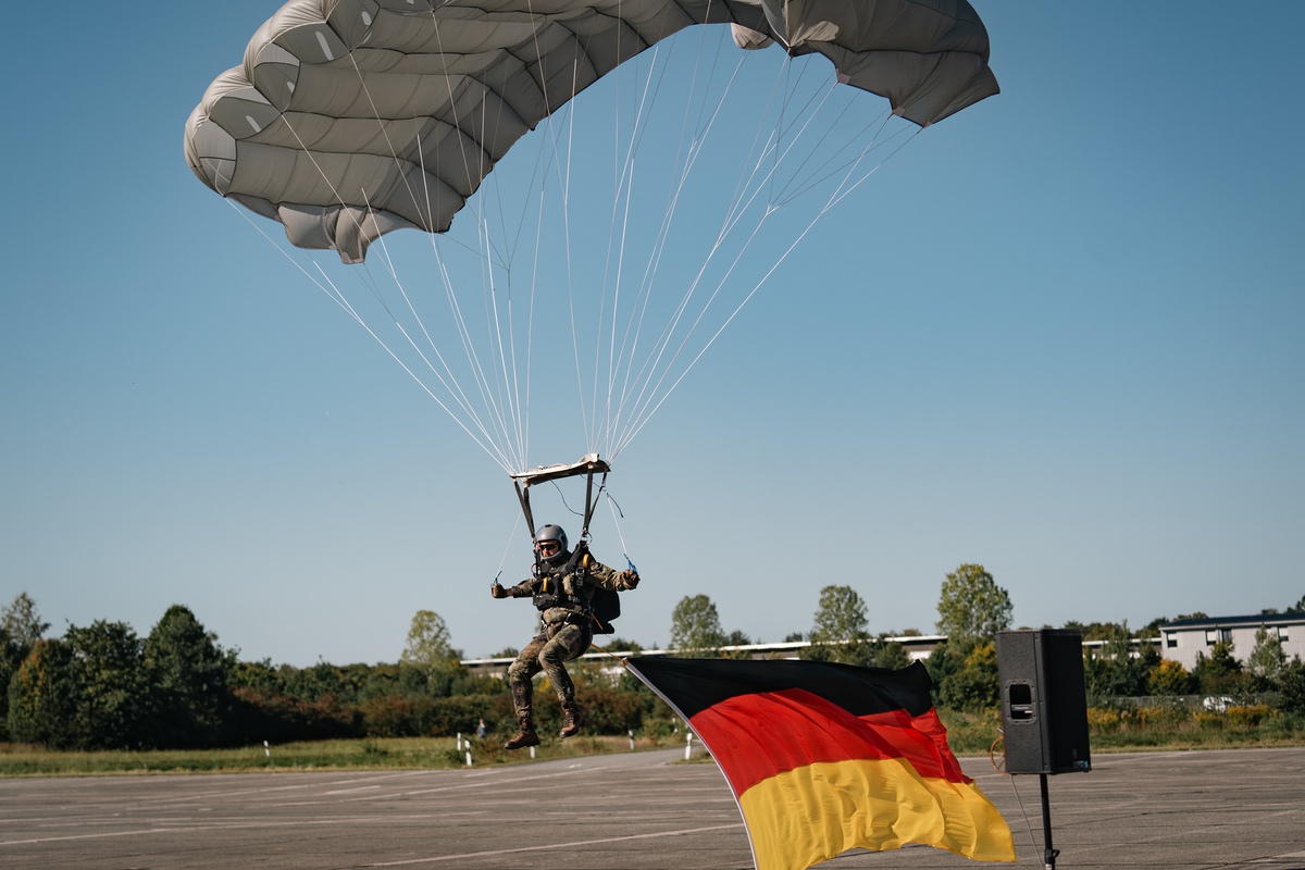 Ein Fallschirmspringer ist im Landeanflug und dem Boden recht nahe. Unter ihm hängt eine Deutschlandflagge.
