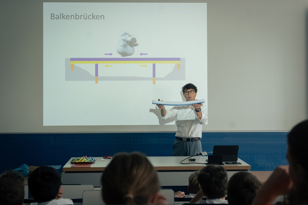 Prof. Braml steht vor einer Präsentation auf der eine Balkenbrücke abgebildert ist. Er hält ein Schaumstoffteil in der Hand und erklärt den Kindern, worauf es beim Brückenbauen ankommt.