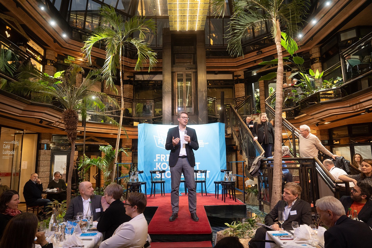 Markus Blume steht auf einem Podest, hinter ihm stehen Stühle vor einem Banner, links und rechts vor ihm sitzen die Zuhörenden an Tischen, neben ihm stehende Zuhörende auf einer Treppe. Um ihn herum stehen Grünpflanzen, er spricht mit einem Mikrofon in der Hand zum Publikum.