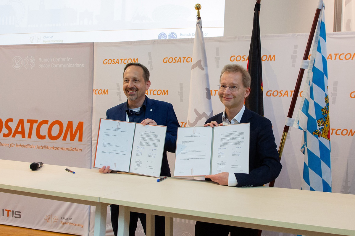 Prof. Knopp und Prof. Huber stehen nebeneinander, beide halten Mappen mit Schriftstücken hoch. Hinter ihnen sind die Logos der Universität sowie der GOSATCOM zu sehen