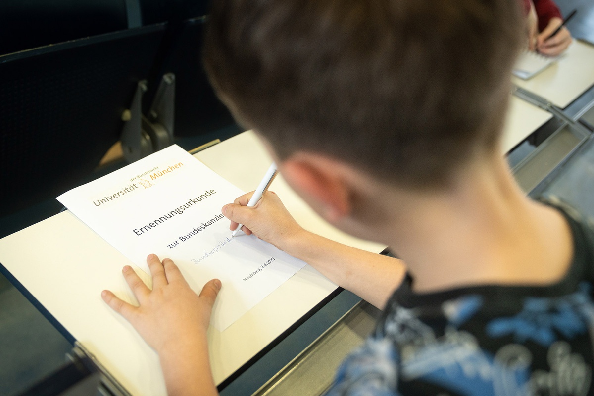 Das Foto wurde über die Schulter eines Kindes aufgenommen, vor sich ein Blatt Papier, das das Logo der Universität zeigt. Darunter steht "Ernennungsurkunde zur Bundeskanzlerin" sowie Ort und Datum. Das Kind schreibt auf dem Blatt Papier, "Bundespräside" kann man bereits lesen.