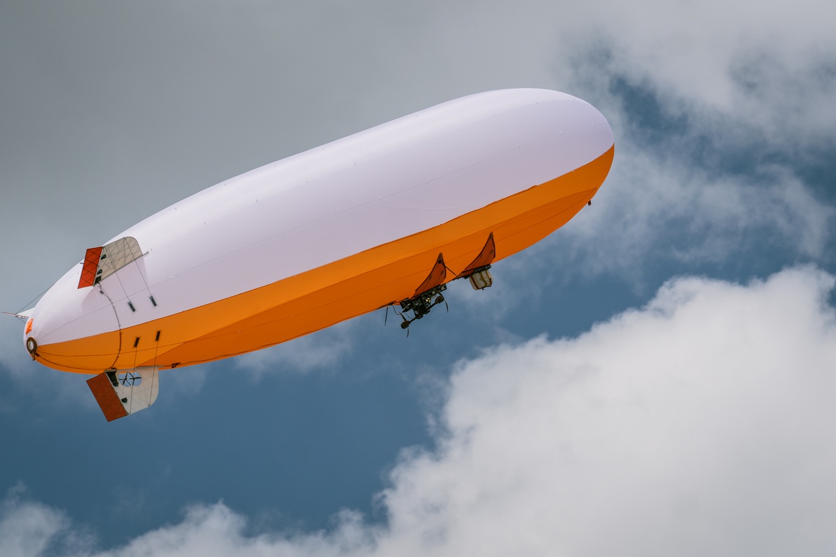 Auf dem Bild sieht man ein orange-weißer Zeppelin im blauen Himmel mit weißen Wolken.