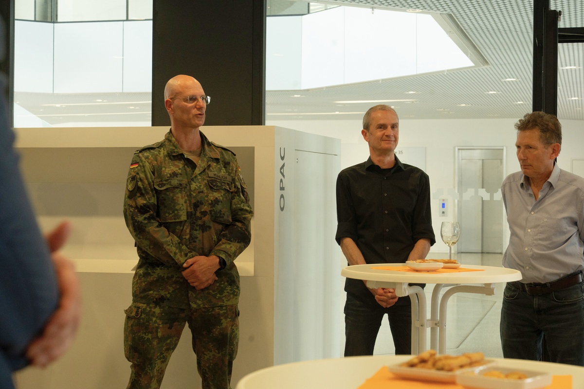 Oberstleutnant Mühlich steht neben zwei Mitarbeitern aus der Bibliothek und teilt seine eigenen Erfahrungen bei der Ausstellungseröffnung