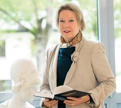 Bild der Präsidentin a. D. Prof. Merith Niehuss, die vor einem Fenster steht und ein aufgeschlagenes Buch in den Händen hält. Neben ihr steht eine Büste der Göttin Athene, Im Hintergrund sind Bäume zu sehen.